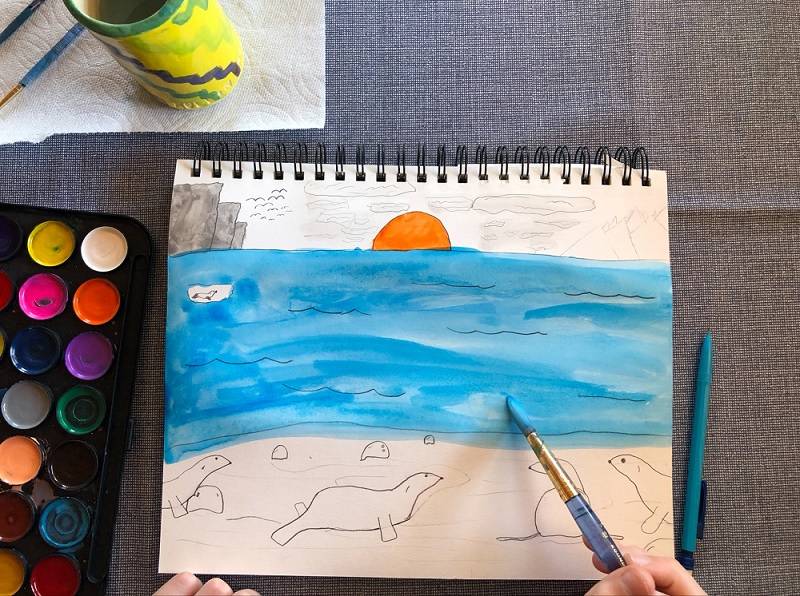 Hand holding a paintbrush, painting a landscape showing seals.