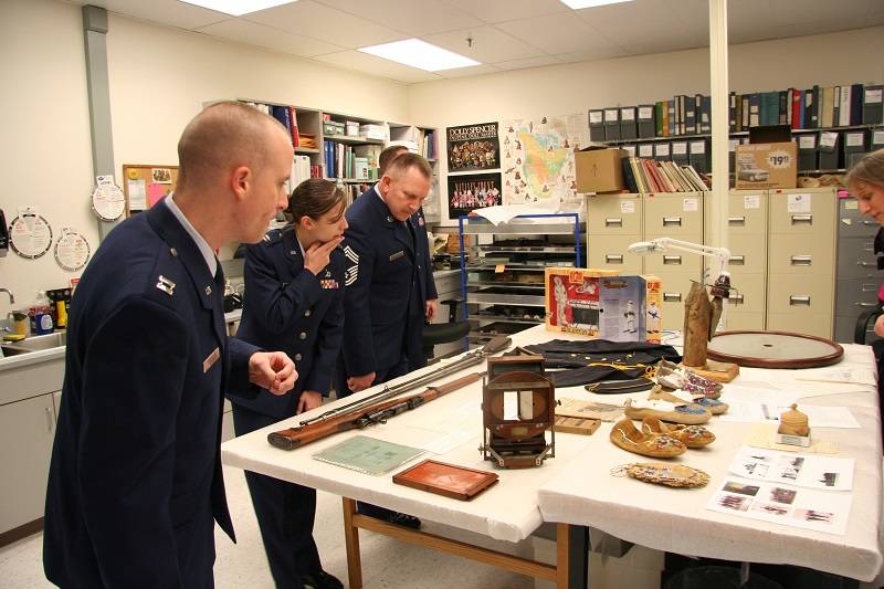 Three people wearing military uniforms looking at a variety of museum objects on a table.