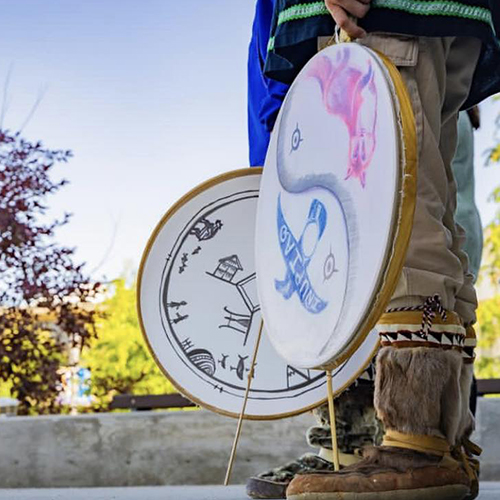 A man in boots is holding Inupiaq drums down by his sides