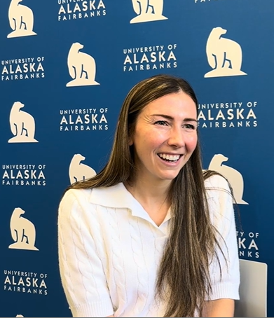 Woman with long brown hair smiles brightly with a "UAF" logo backdrop in blue and white