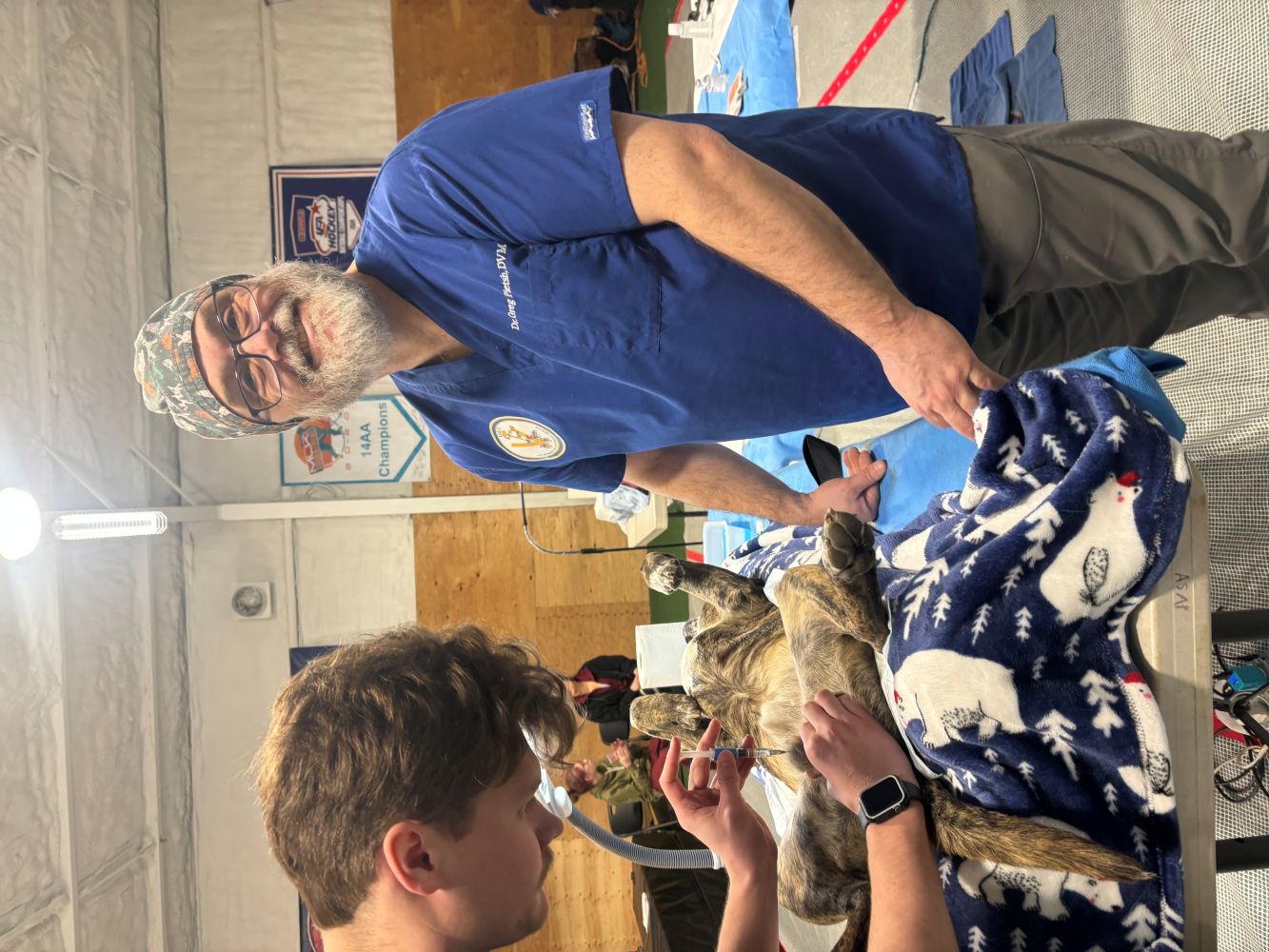 A veterinarian in a blue scrub top smiles at someone off frame with a dog laid out on a table while another person performs a procedure on it in a makeshift clinic setting. 