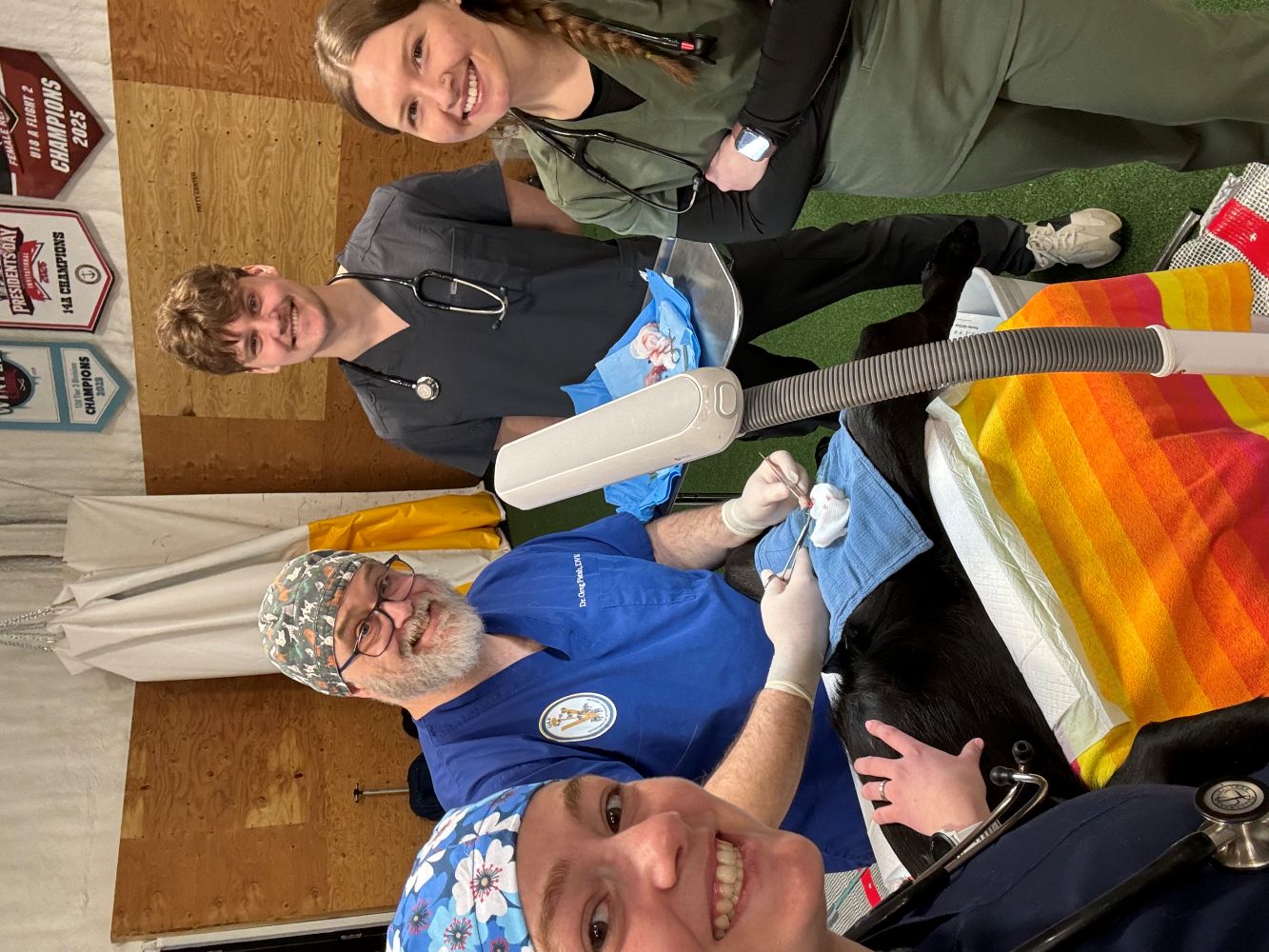 Four people in scrubs, two of who have stethoscopes around their necks, smile for the camera. There is a dog on a table they are surrounding who is being prepared for a procdeure. 
