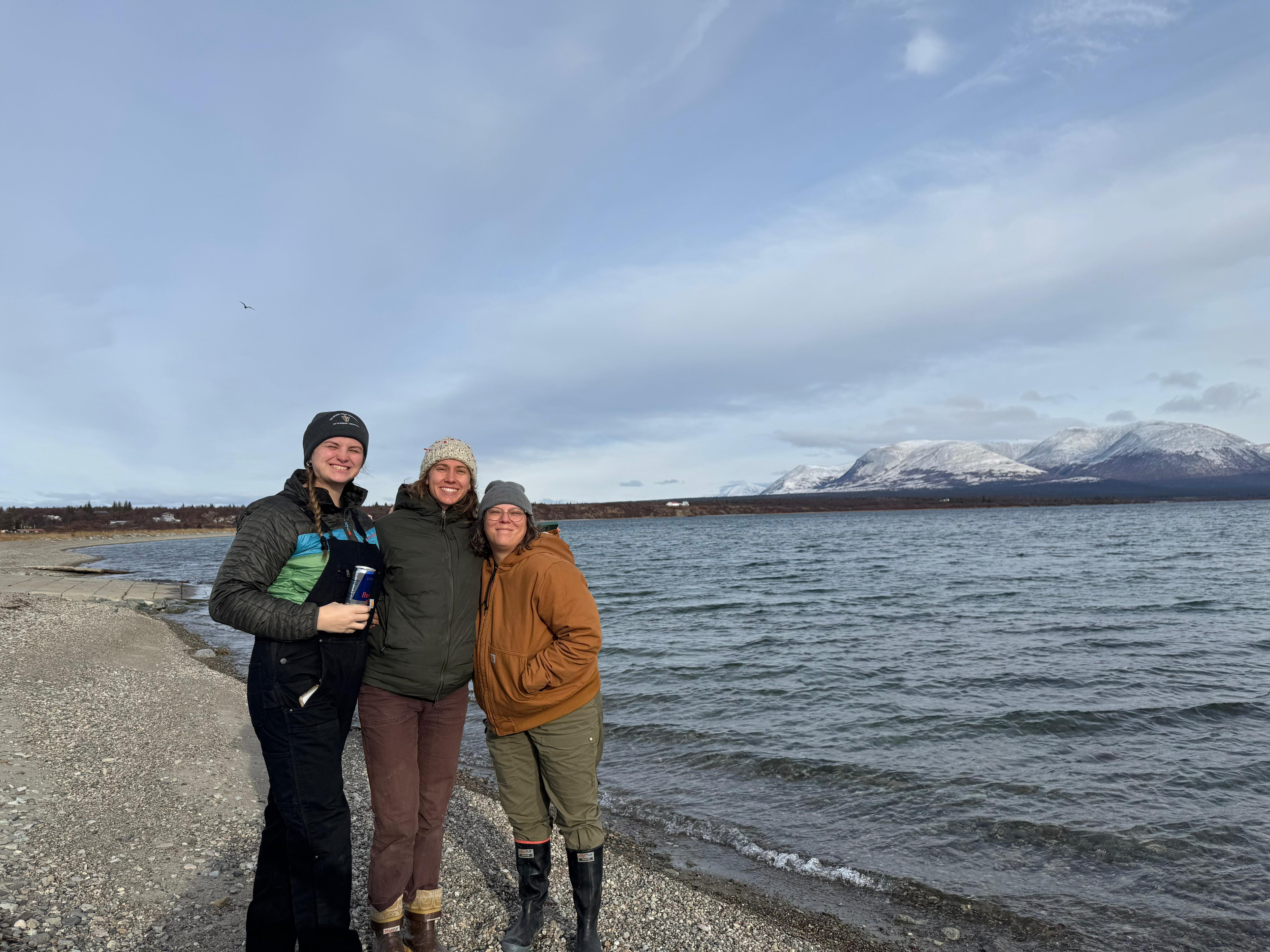 Three women stand smiling together on a pebbly beach with a wide body of water behind them. They are dressed in warm outdoor gear, including jackets, beanies, and boots. In the background, snow-capped mountains rise under a vast, overcast sky.