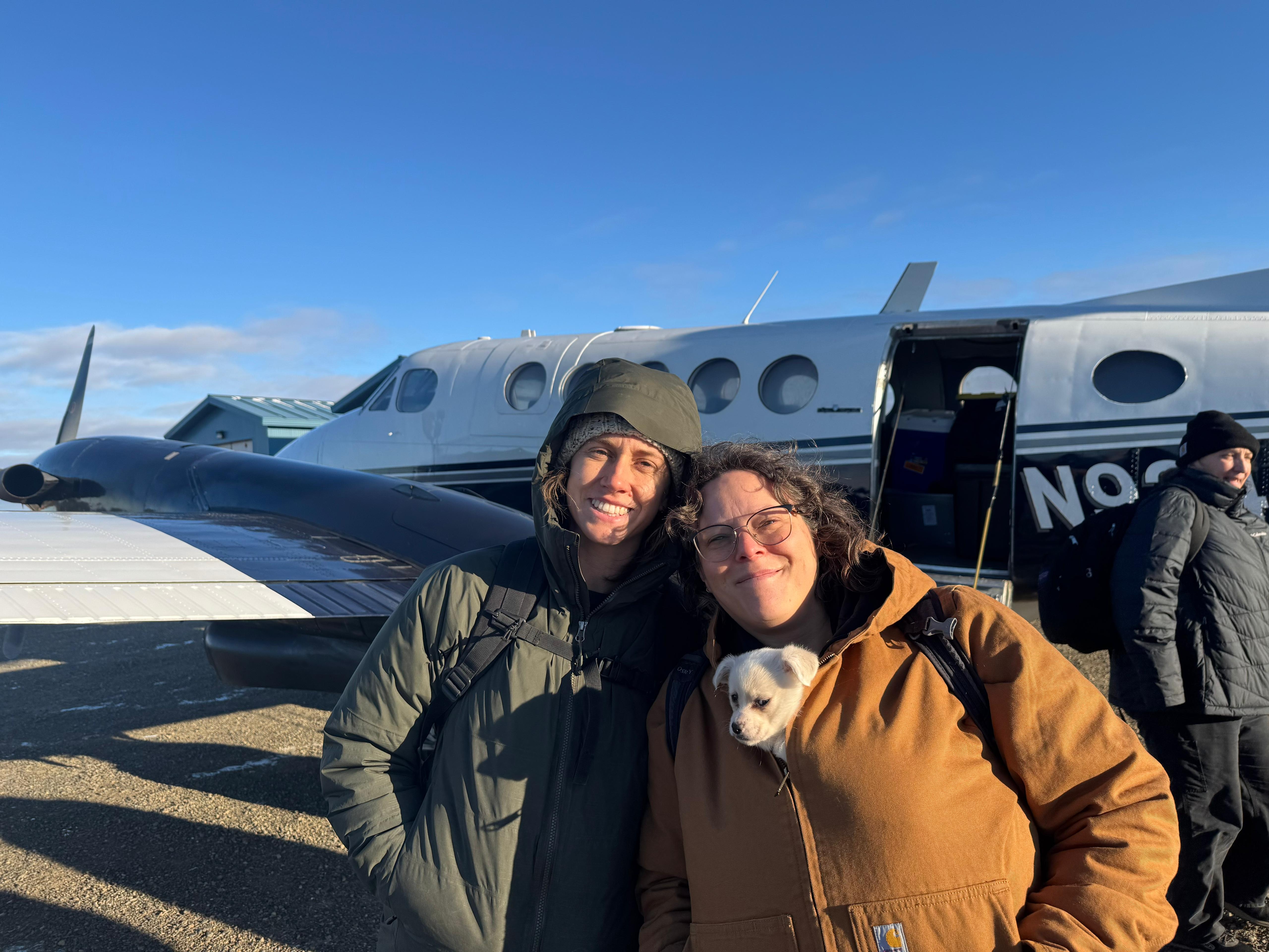 Two women stand smiling in front of a small white airplane on a sunny day. One woman, wearing a brown jacket, has a small white puppy tucked inside the front of her coat. The other woman wears a dark green hooded jacket. In the background, another person walks near the plane's open door under a clear blue sky.