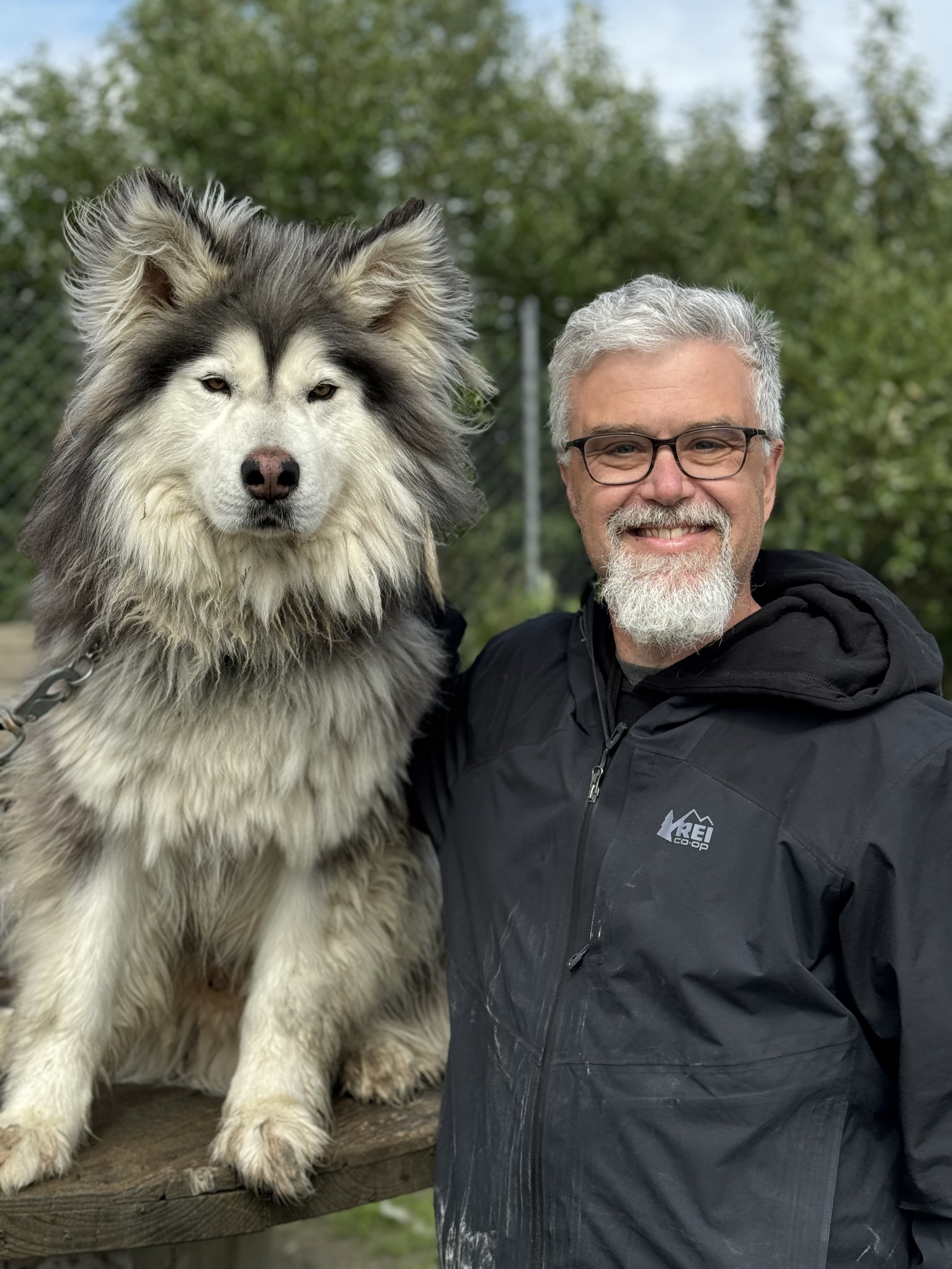 A man with grey hair, beard, and glasses poses next to a large shaggy haired husky on a summer day with trees in the background.