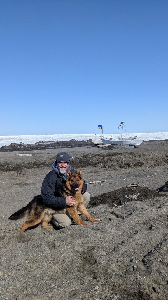 A bearded man wearing a coat and beanie holds a large german shepherd on a black sand beach with a shored white boat in the background