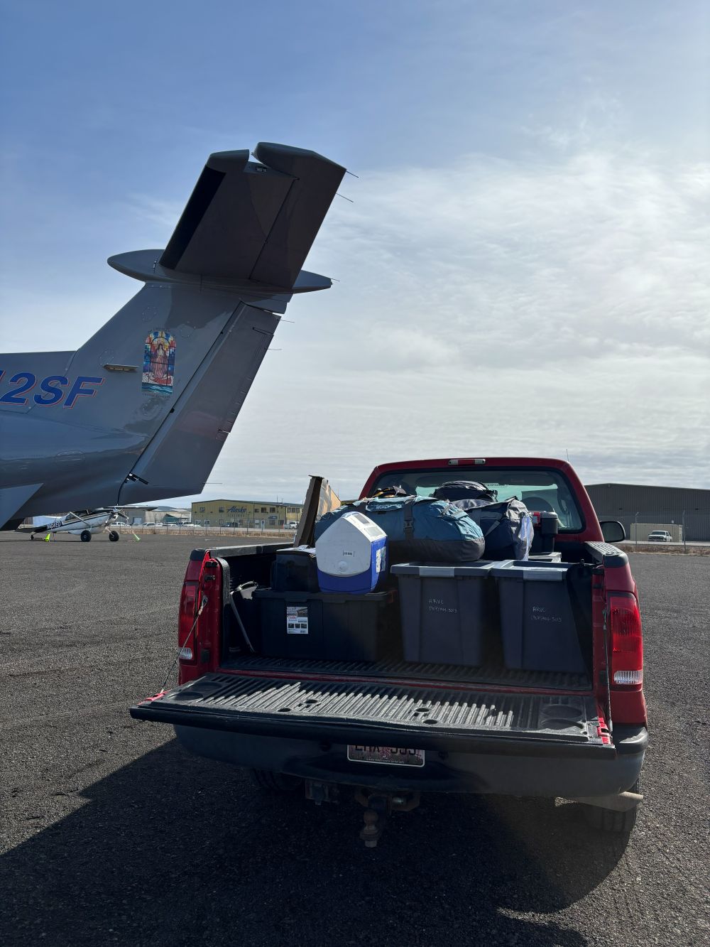 A pick up truck on the tarmac of an airport is filled with tubs and coolers of medical supplies with the tail of a plane in the background on a sunny day
