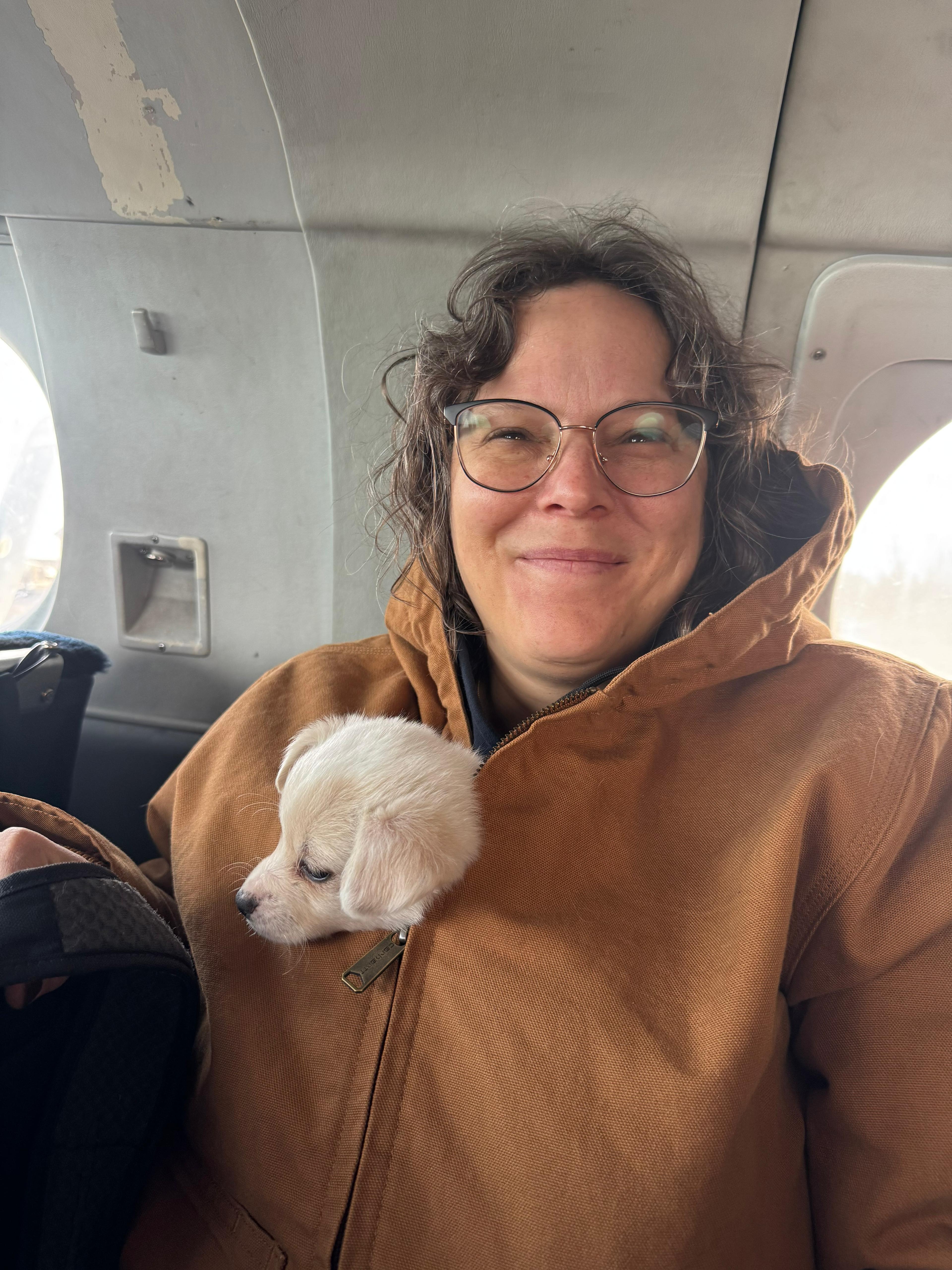 A woman with short brown hair, glasses, and a brown coat smiles with a small white puppy snuggled in her coat