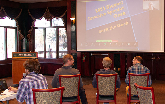 A row of people in a conference room sit in front of a large screen that says 2024 Biggest Invasive Species Geek