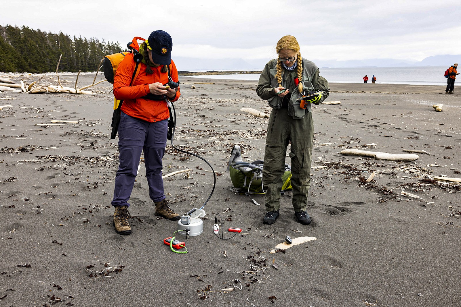 Two female scientists in outdoor clothing and loaded with gear stand on a beach examining small data-collecting devices.