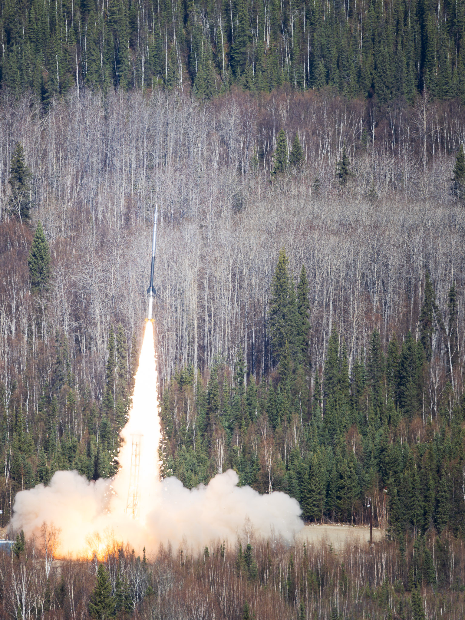 A NASA sounding rocket carrying the FOXSI-5 mission lifts off from Poker Flat Research Range north of Fairbanks at 11:23 a.m. Thursday, May 14, 2026. A bright plume of flame and smoke rises from the launch pad as the rocket climbs above a dense forest of spruce and leafless birch trees. The FOXSI-5 mission is part of a continuing NASA-led program to study the sun.