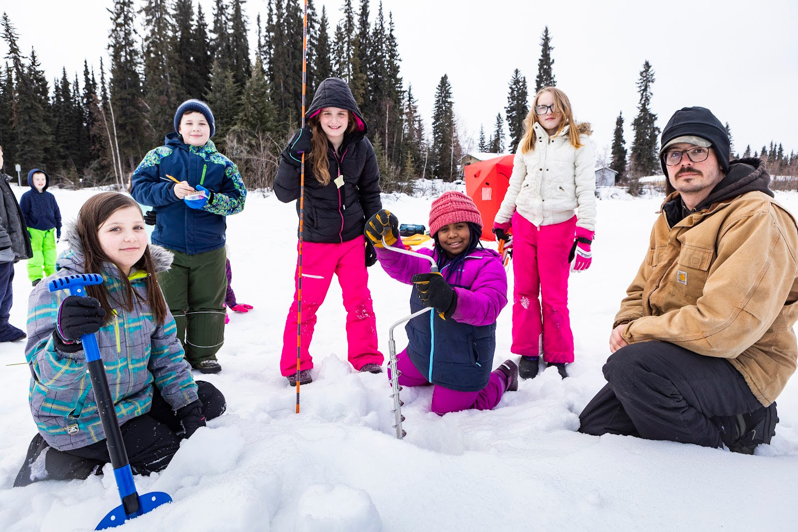 An adult and a group of children in colorful clothing drill a hole in the snow and ice.