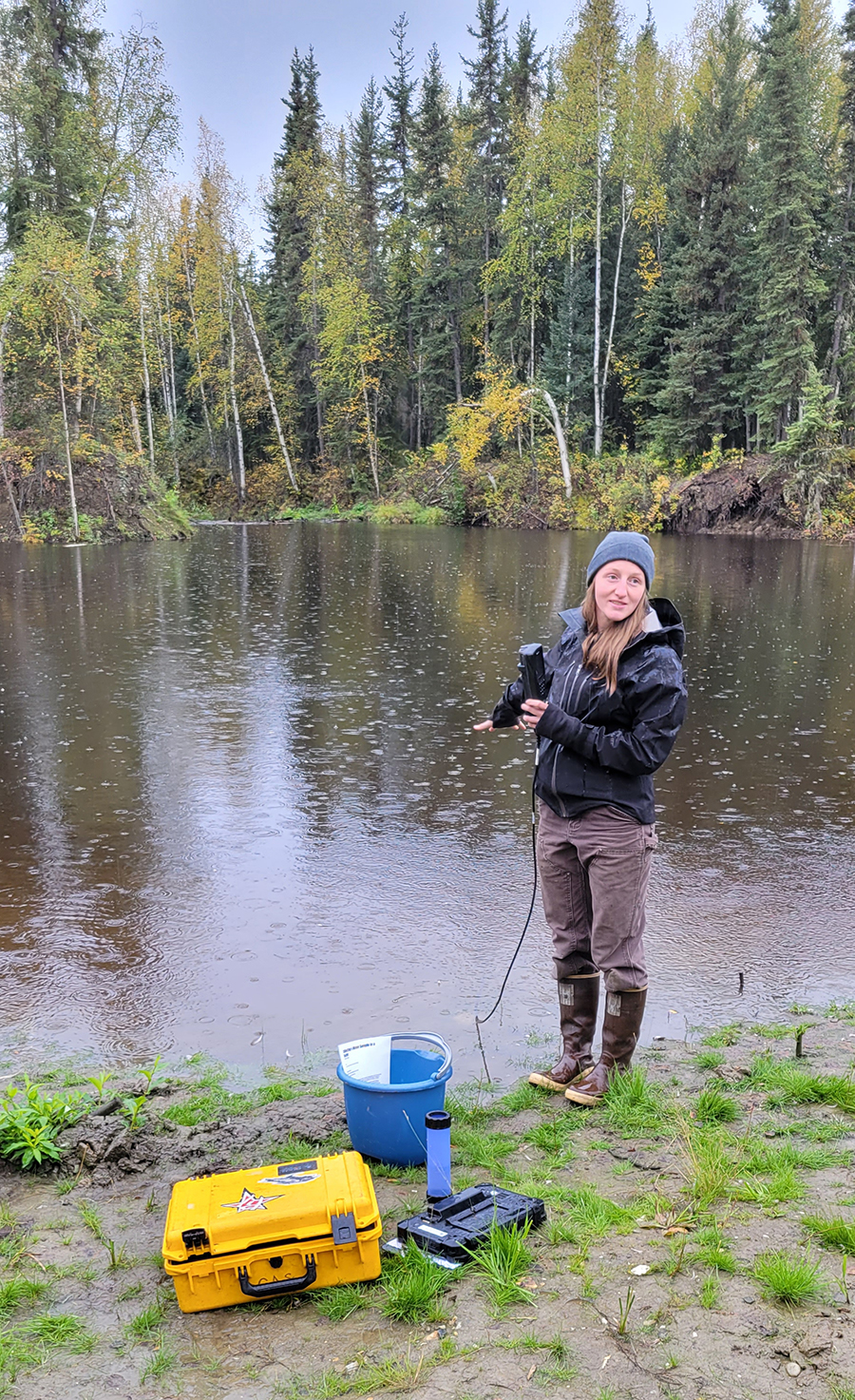 A woman in a jacket and knit cap stands near a body of water with water quality testing equipment in a bright yellow box. 