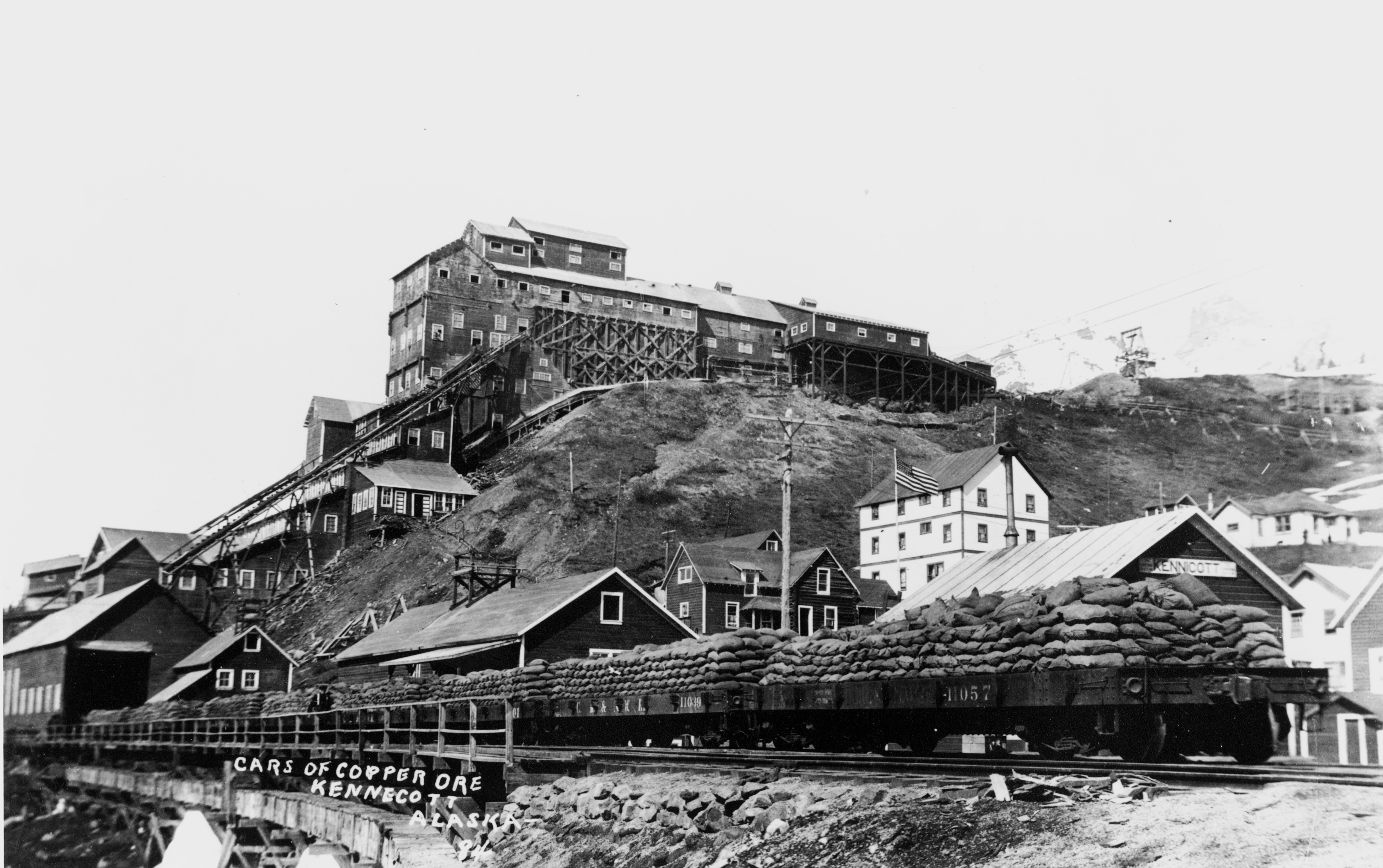 Black-and-white historic photo of Kennecott Mines, with railcars piled high with copper ore awaiting shipment on the Copper River and Northwestern Railway. Mine buildings, conveyors and other structures rise on the hillside behind the train.