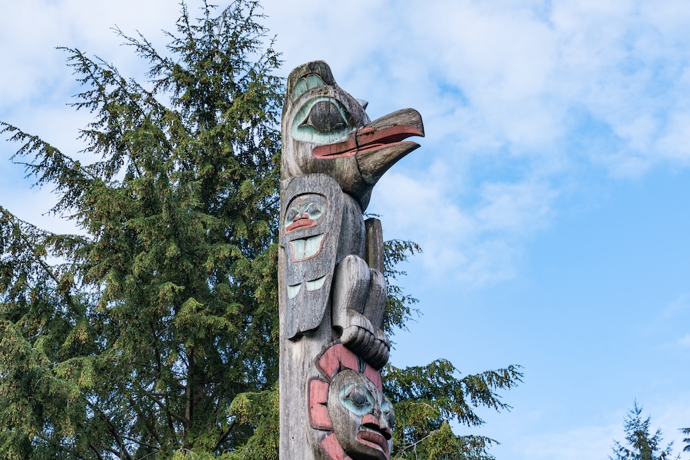 A faded totem pole with a red-beaked bird at the top in front of an evergreen tree.