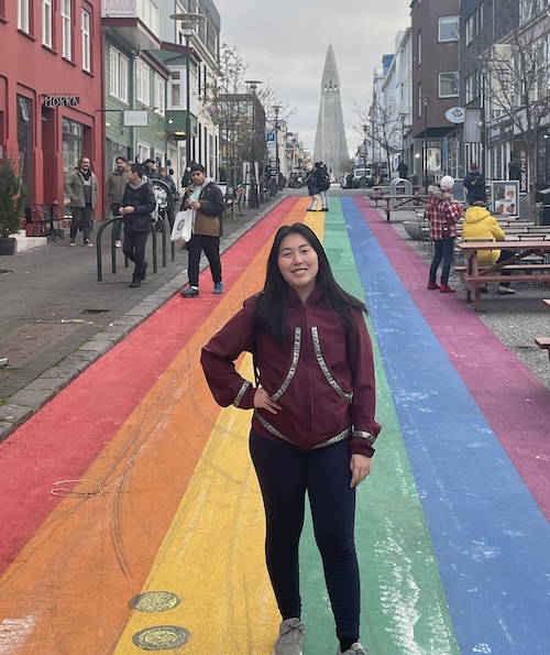 A young hispanic woman stands on a street painted in rainbow colors.