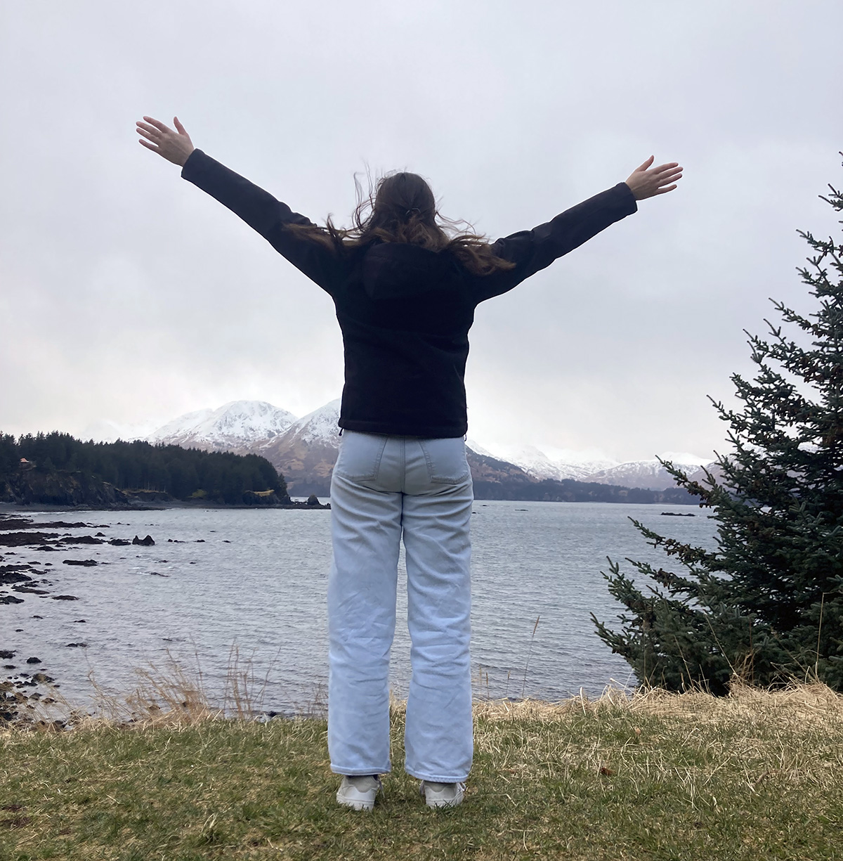 A woman stands on the waterfront with her arms stretched out