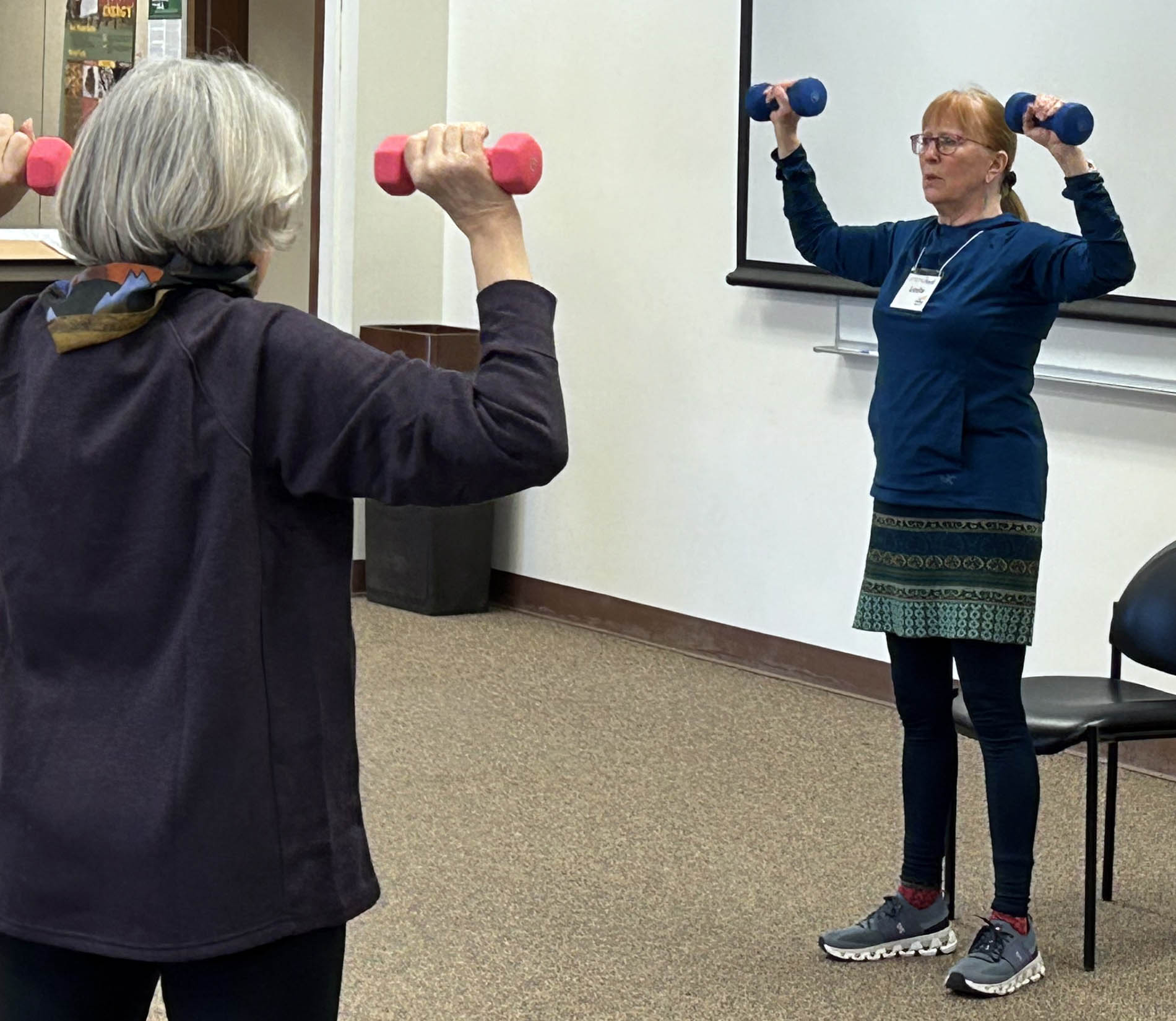 A woman at the front of a classroom lifts hand weights as she shows an older woman an exercise.