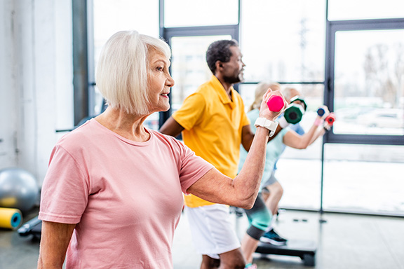 A woman with white hair and a pink shirt holds a hand weight in an exercise class.