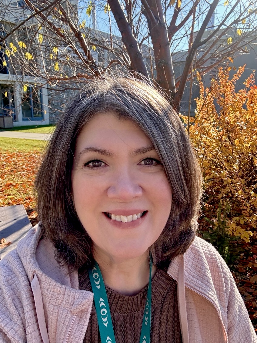 A smiling woman looks into the camera in front of bushes with brightly colored leaves and a university building