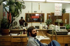 A busy barbershop with a man sitting in the forground with a person taking a picture visible in the mirrors.a