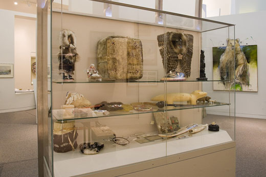A display case has many Alaskan Native items with a painting of a bear in the background.