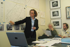 A woman points at a schematic on the wall while two men look on in a conference room.