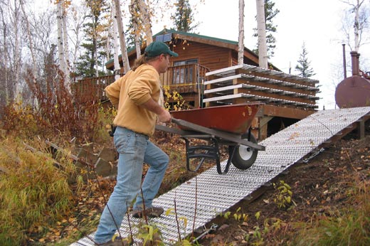 A man pushing a wheelbarrow up a aluminum grate ramp, with a building in the background.