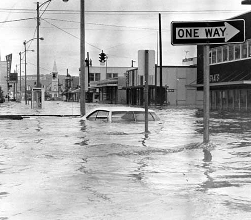 Close-up view of flooded street, with car under water up to windshield; stores and shops border both sides, and a church is in background.