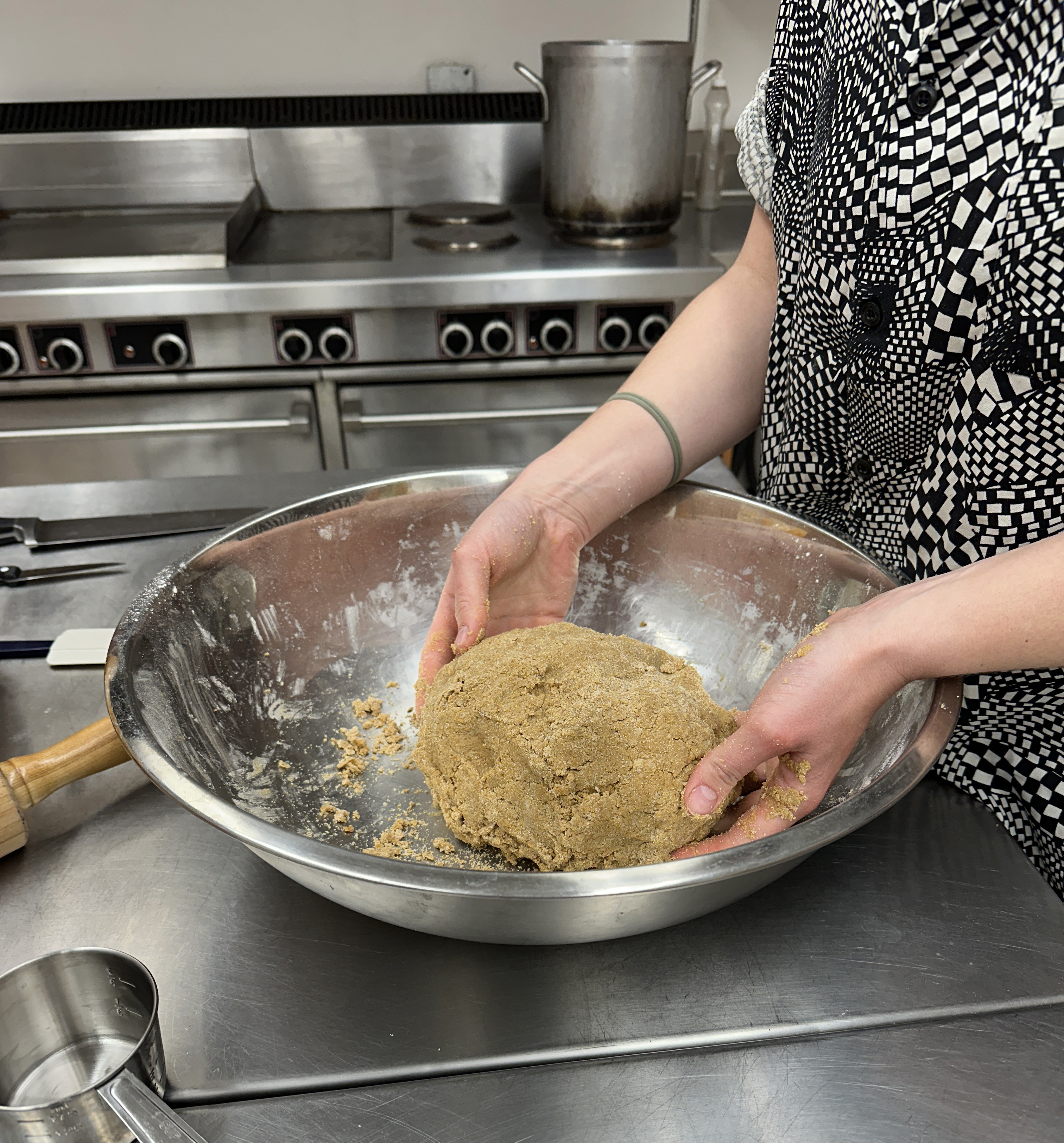 A person shapes a ball of barley cracker dough in a metal bowl. A large stove and pot are in the background.