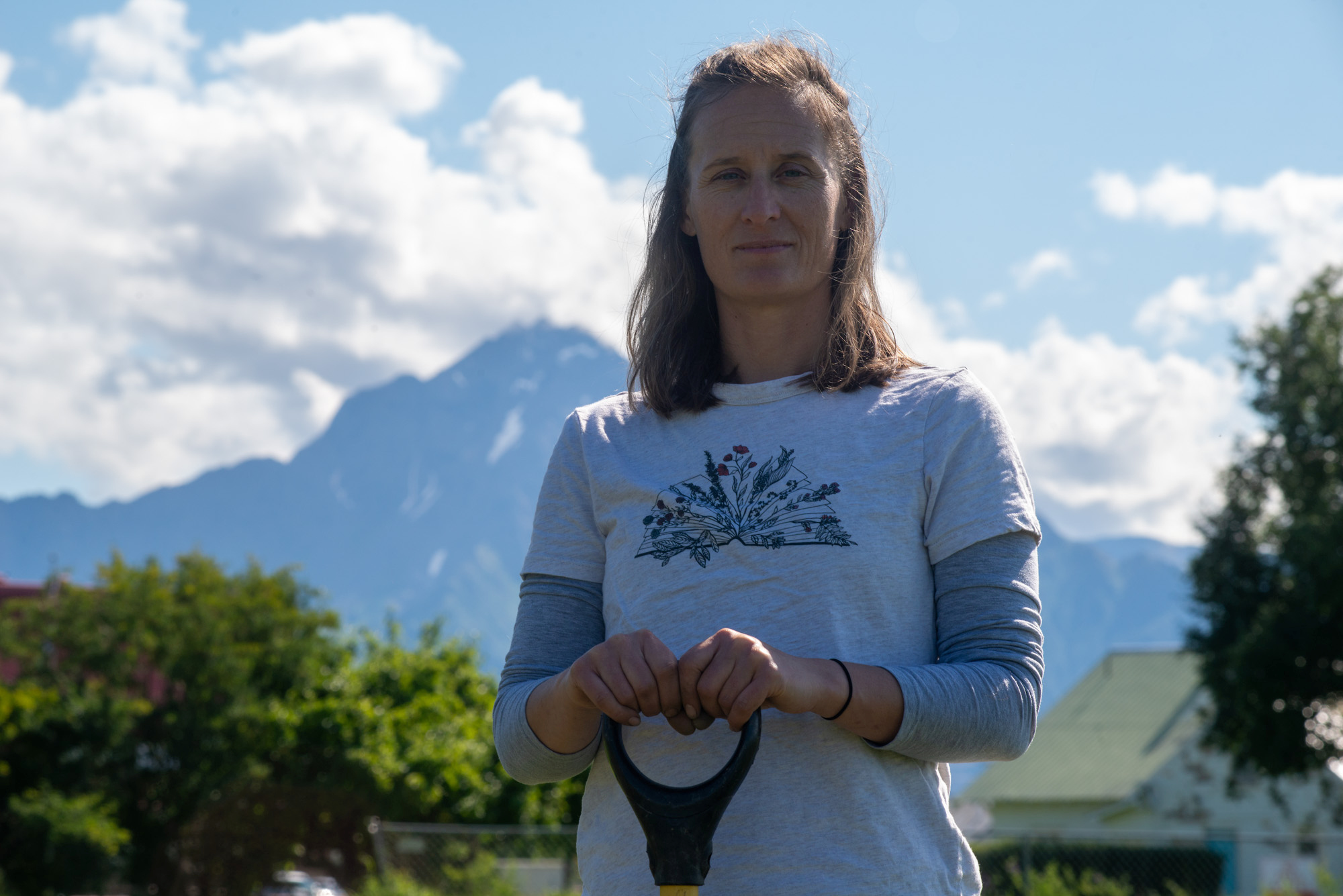 A woman, Caley Gasch, stands in front of snow-capped mountain with a shovel in her hands