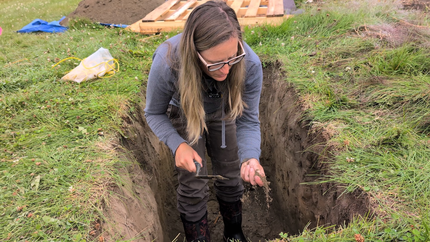 A woman crumbles soil in her hand while standing in a pit in an agricultural field.