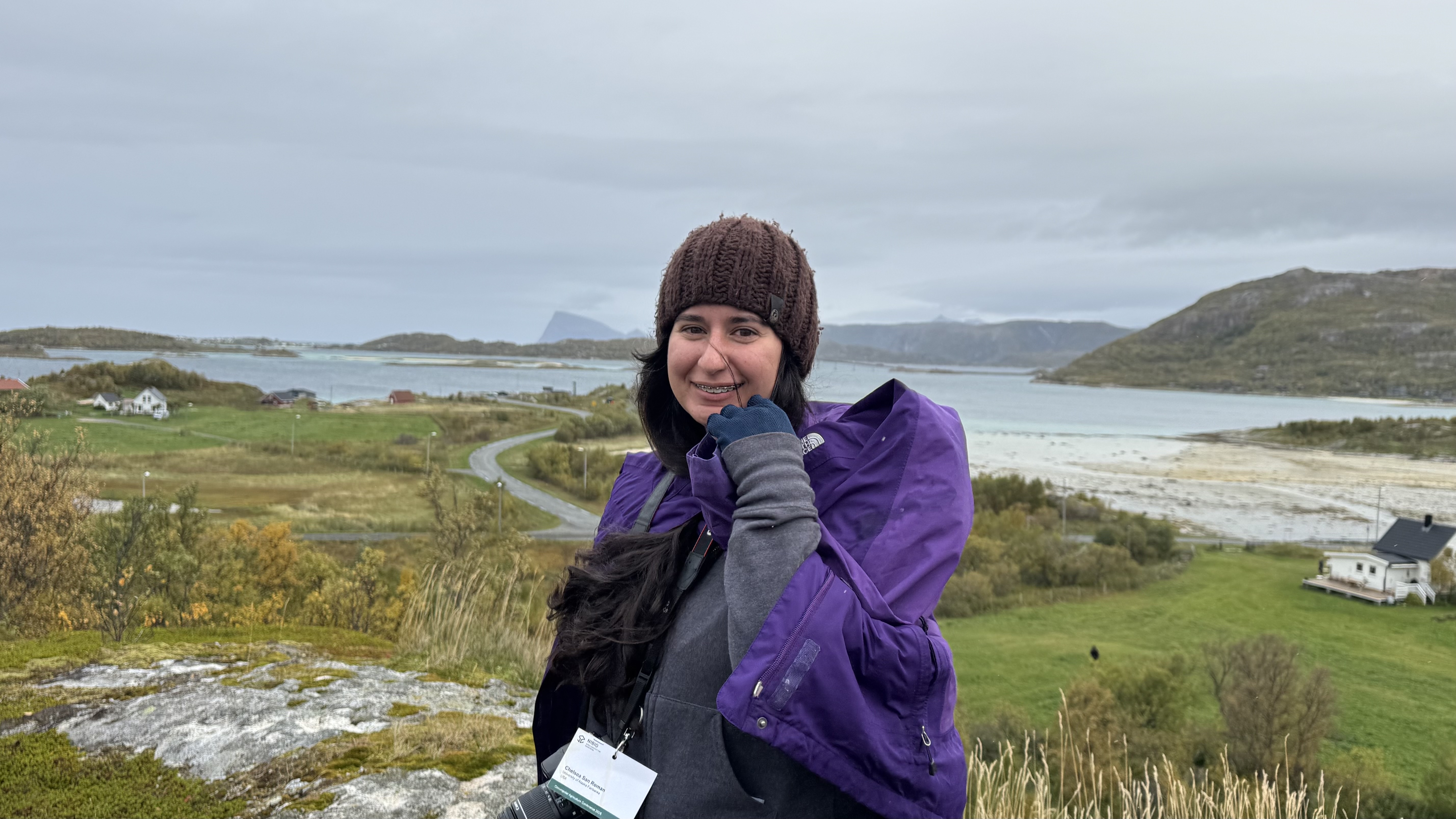 A woman in a gray and purple parka and brown knitted cap stands in front of farmland backed by a bay with mountains in the background