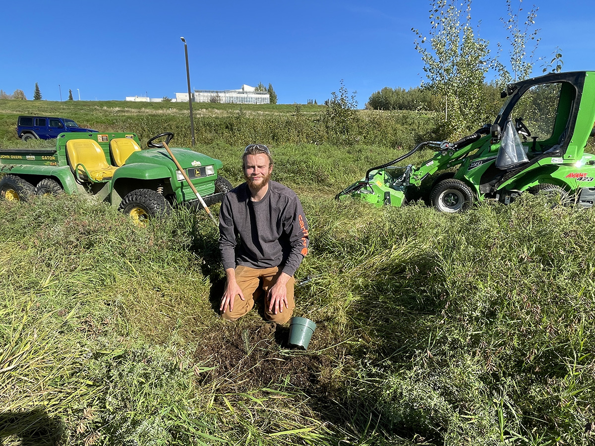 A man kneels on the ground next to freshly planted saplings, flanked by farm equipment.