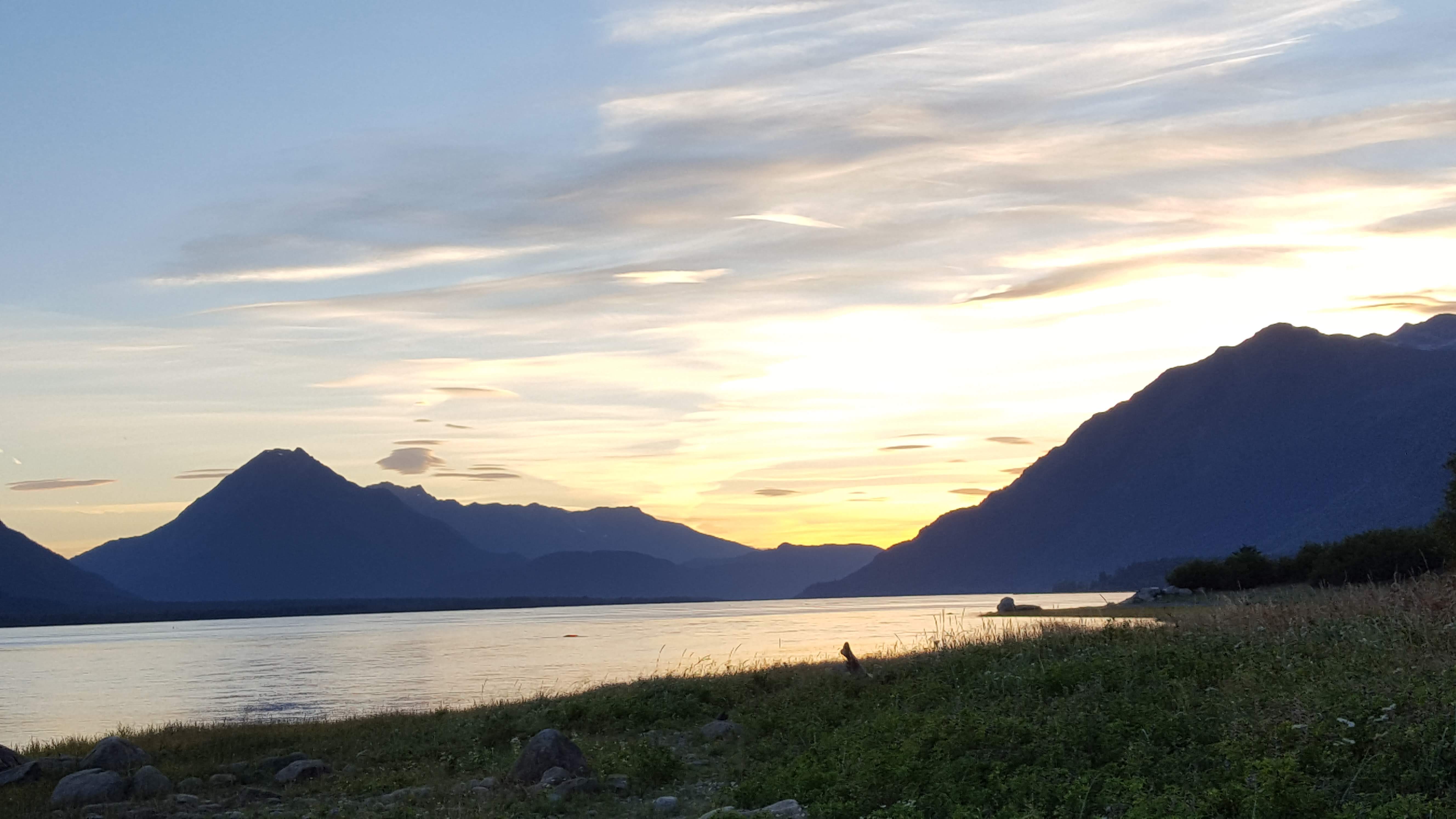 A river runs toward mountains that are silhouetted by the sun and clouds