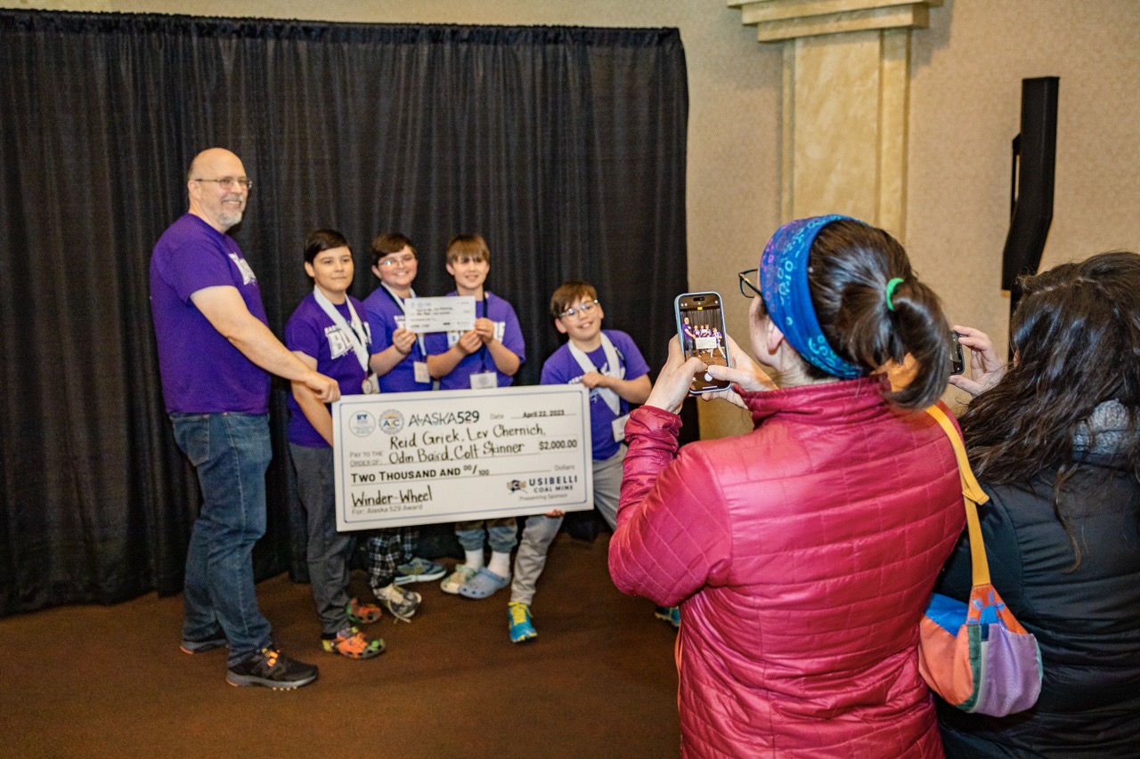 A man and four middle school students, all wearing purple t-shirts, hold a large presentation check. A woman in the foreground is taking their photo with a phone camera.