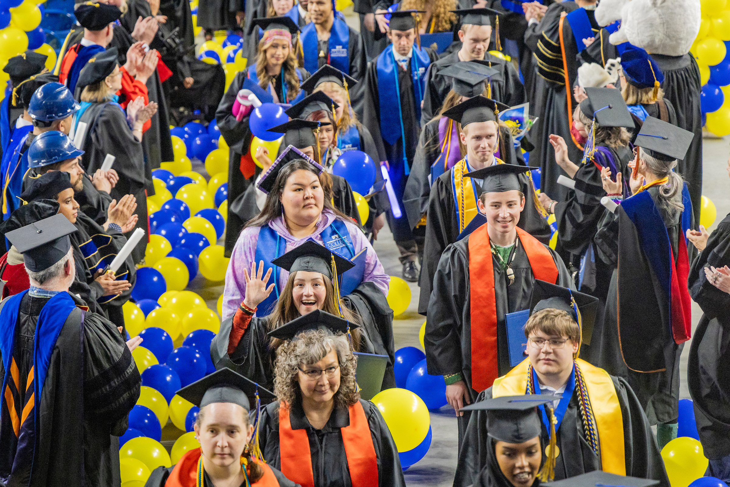 At the 2025 commencement ceremony, a crowd of UAF graduates in caps and gowns process out of the Carlson Center amid blue and gold balloons, smiling and waving, as faculty and staff applaud from the sides.