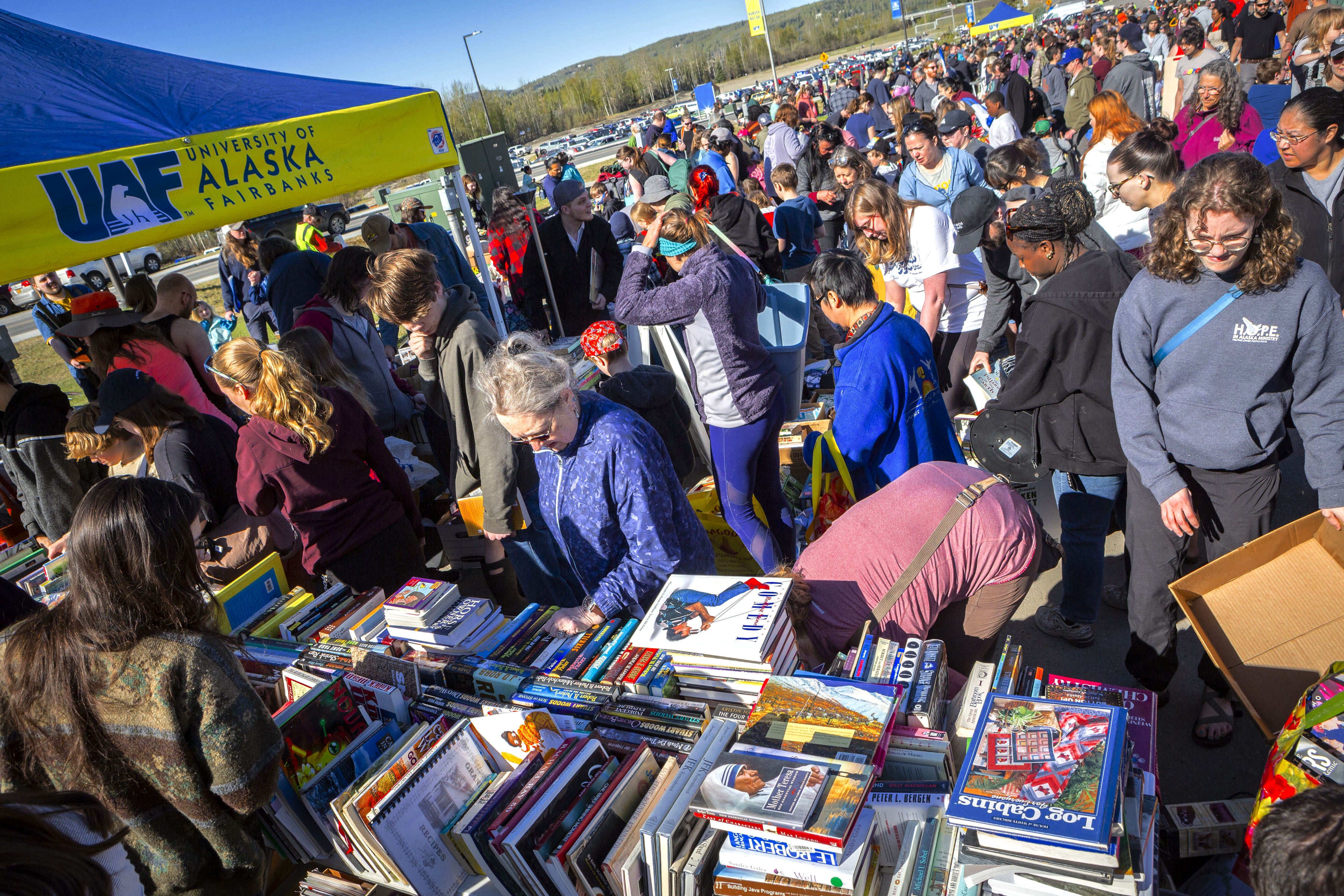 A large crowd browses tables piled high with free books and other items at the 2025 Really Free Market outdoor recycling event, with a UAF-branded tent visible in the background under a clear blue sky.