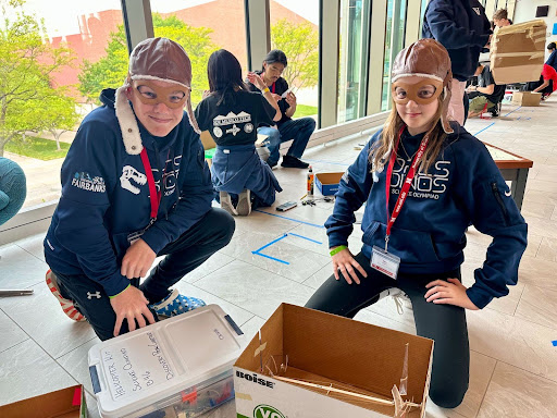 Two middle school students squat next to boxes with supplies for the helicopter challenge. They are wearing team sweatshirts, name tag lanyards, and aviator hats. Students from other olympiad teams are behind them in the hallway.