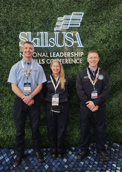 Instructor Jamie Bero stands between UAF student firefighters Tyler Christensen and Daniel Hayes smiling and wearing medals in front of a SkillsUSA backdrop.
