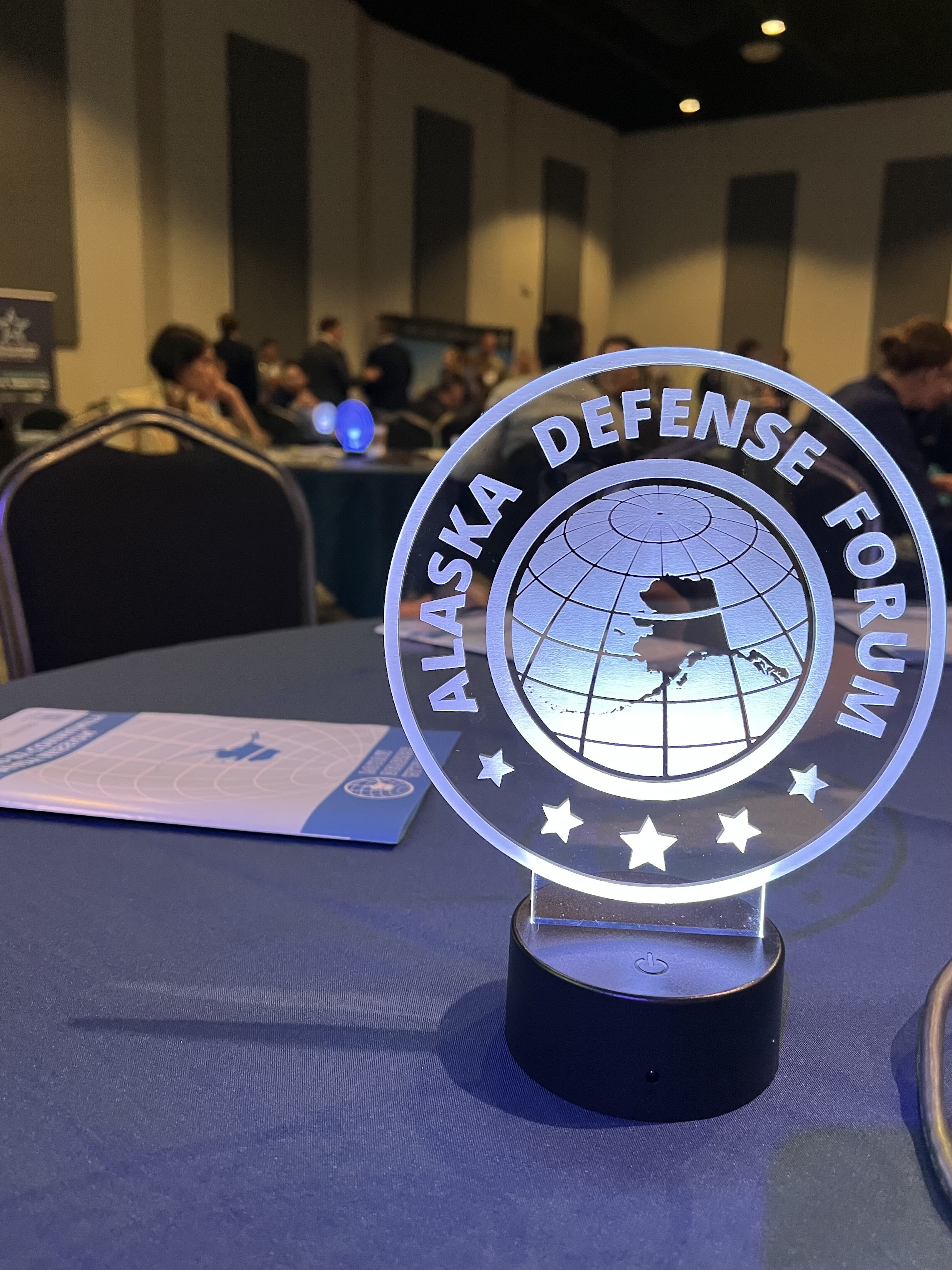 A glowing Alaska Defense Forum sign sits on a table at the 2025 event at the 8 Star Event Center in Fairbanks. The circular sign features an outline of Alaska over a globe, with conference attendees visible in the background.