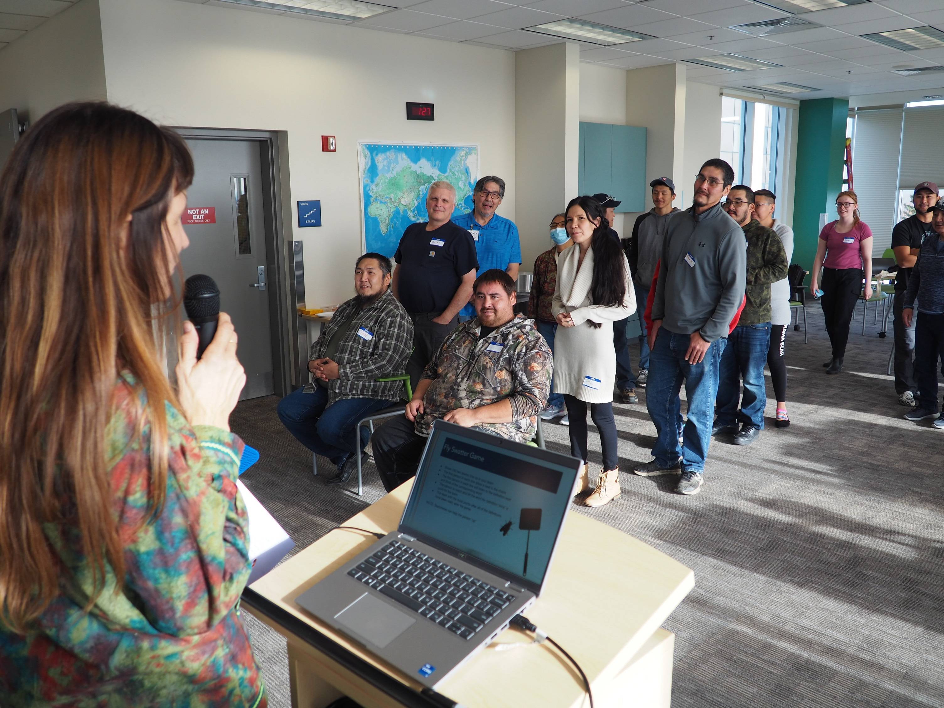 A woman stands in front of a computer speaking into a microphone in a room of people.