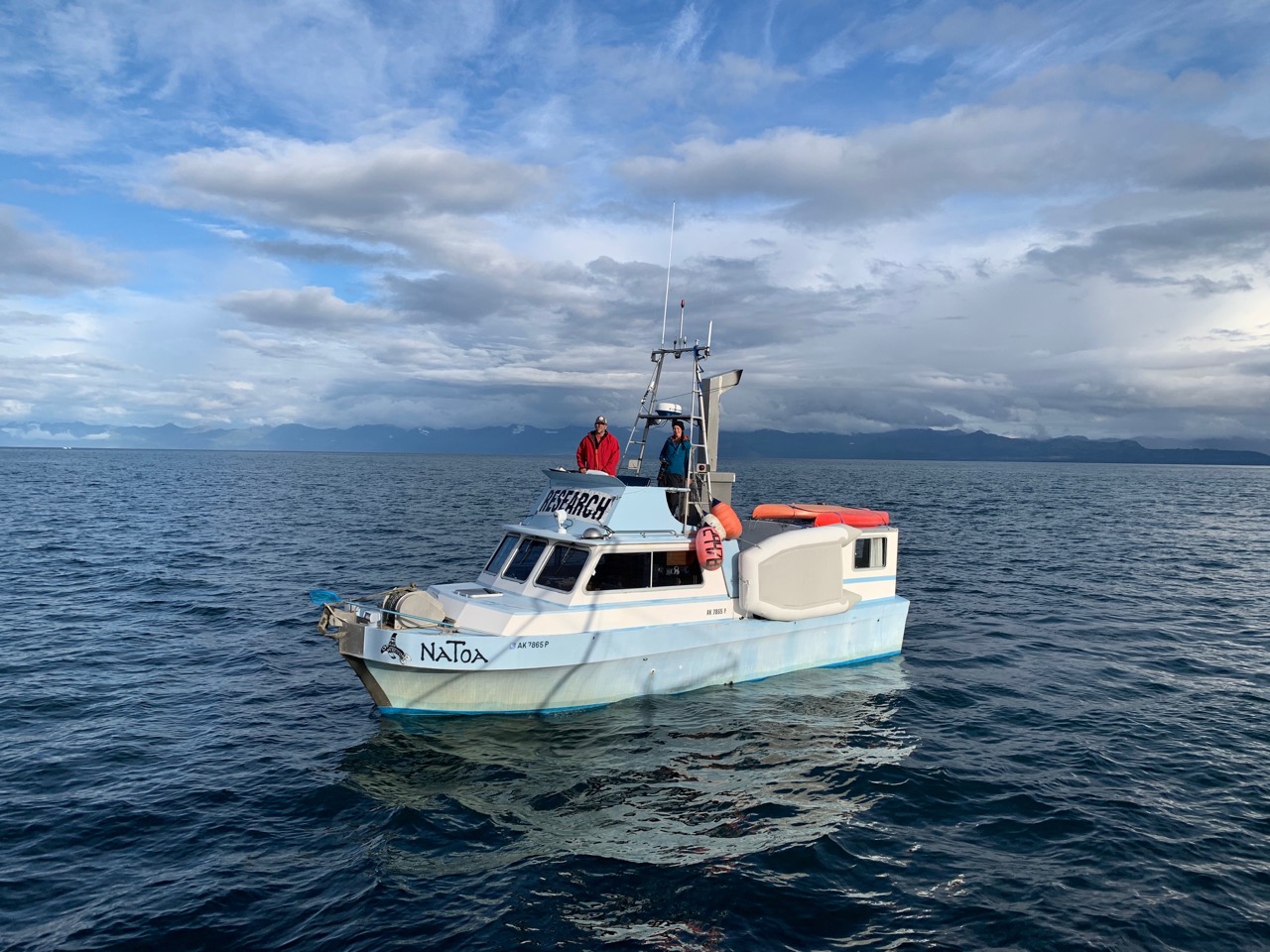 A white and light blue 32-foot boat, Natoa, sits on ocean waters dappled with small waves. A woman and a man stand on an open bridge atop the fore cabin, under a framework holding antennas and a compact radar dome. An inflatable life raft hangs on the side of the boat. An orange kayak and a red kayak sit atop the flat roof of the rear cabin. Dark blue mountainous land forms the distant background. 