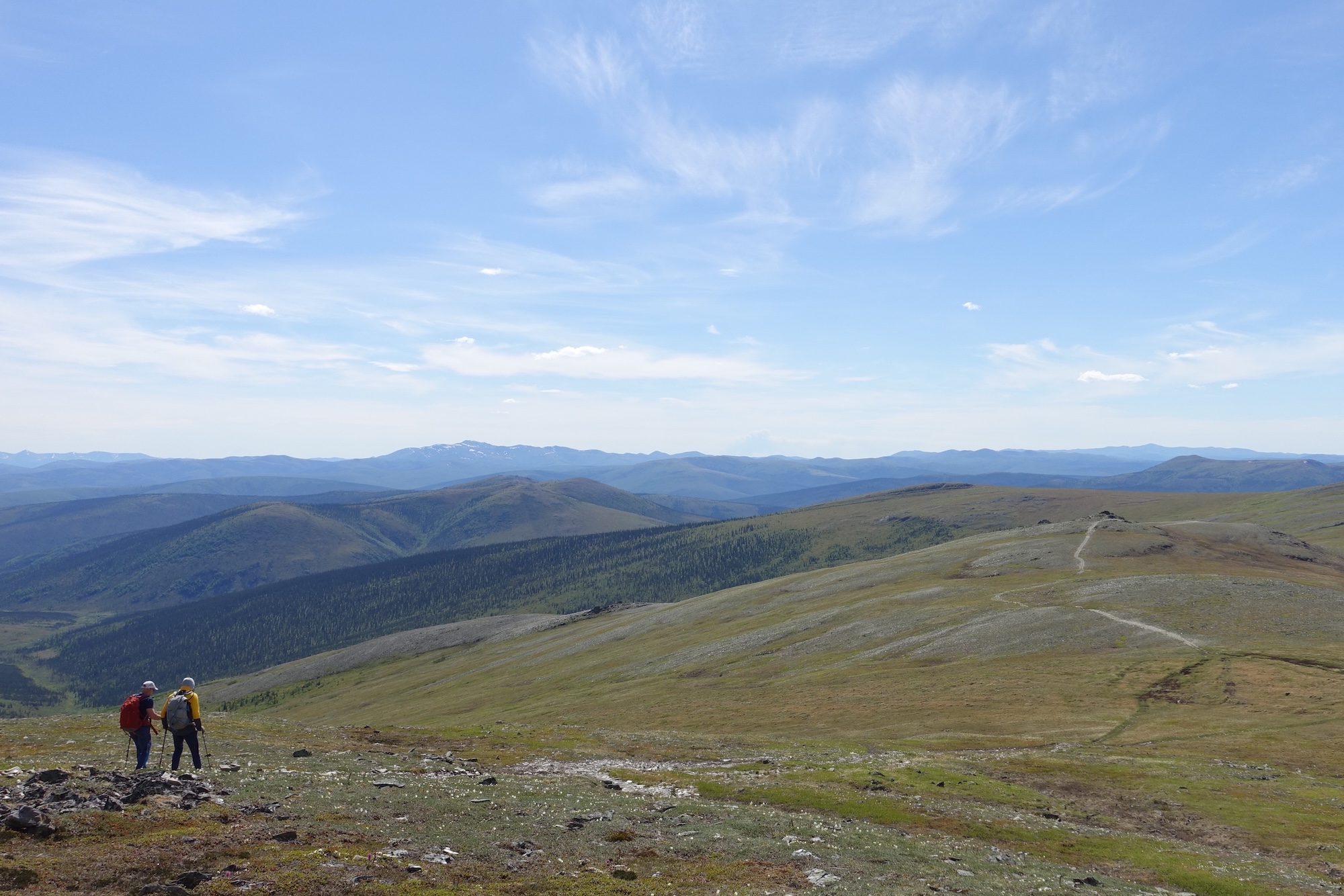 On a sunny day with a few wispy clouds, two people hike on a trail over a tundra-covered ridge with rolling hills and mountains in the background.