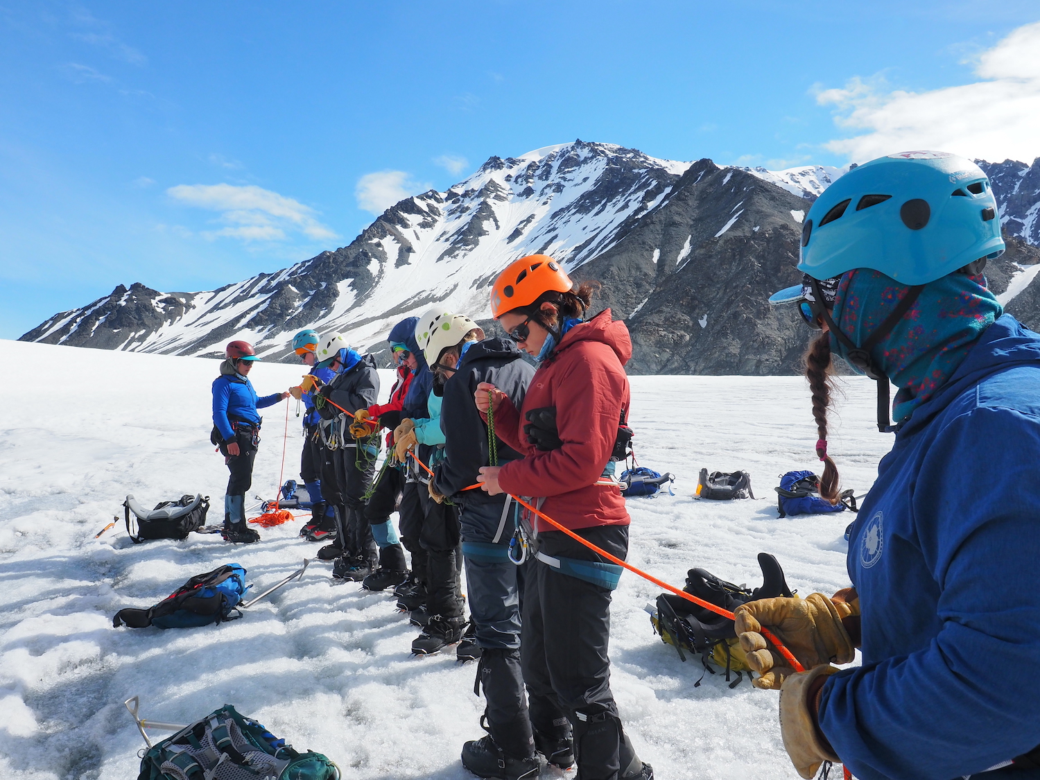 Girls in mountaineering gear stand on a glacier while tying ropes to a long rope.