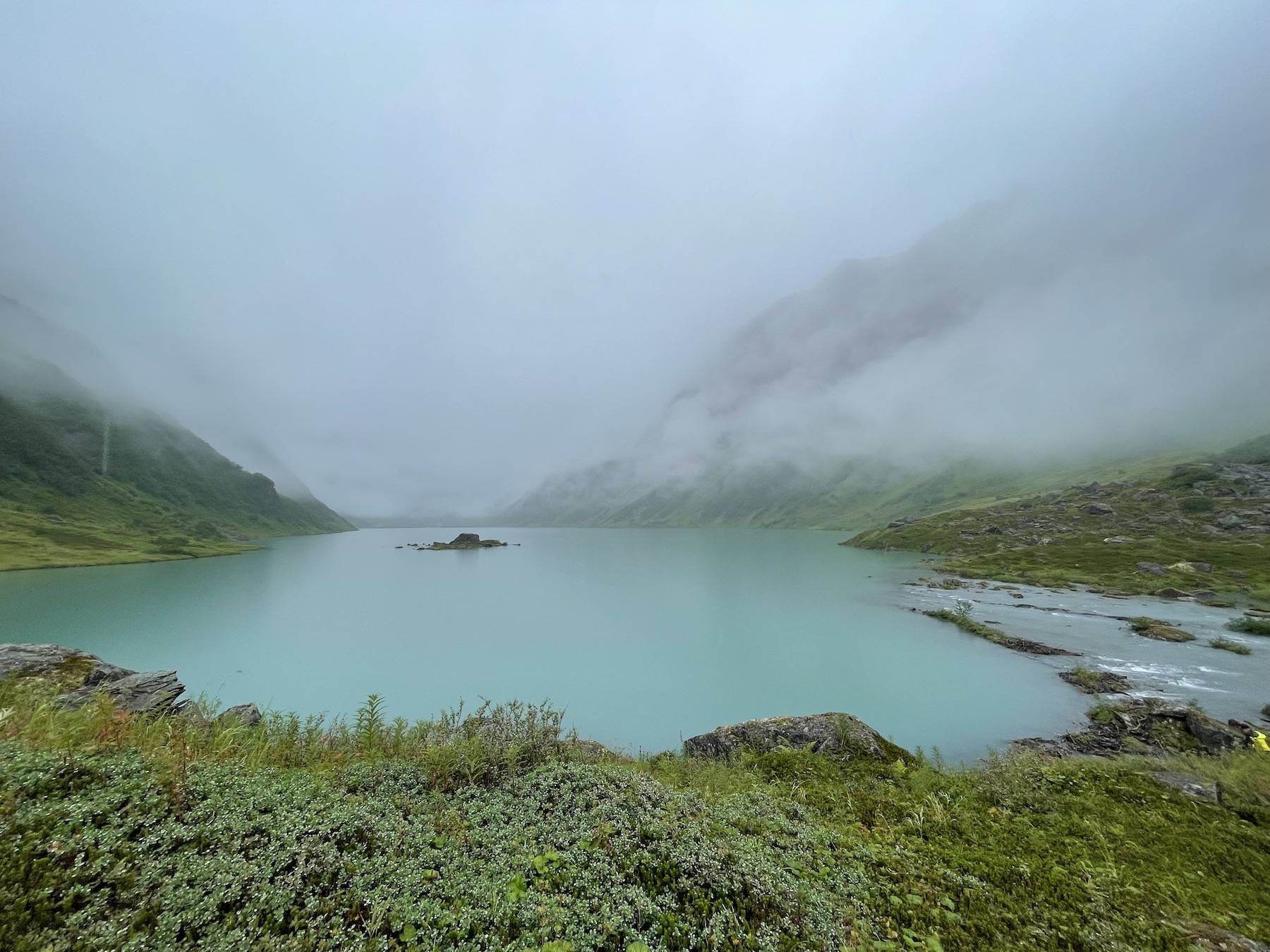 A lake with a small rocky island in the center sits beneath green alpine hillsides and foggy clouds.