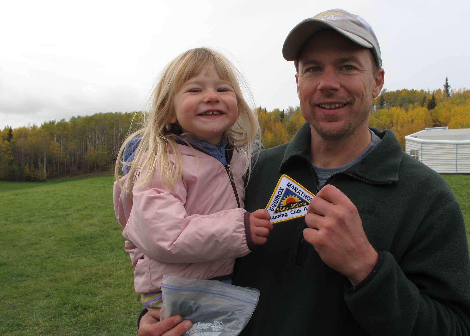 A man in a ball cap holding a young blond girl both hold an Equinox Marathon patch. 