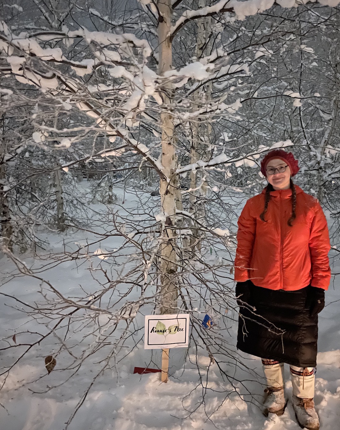 A woman in a red winter jacket and hat stands next to a birch tree in a snowy forest. 