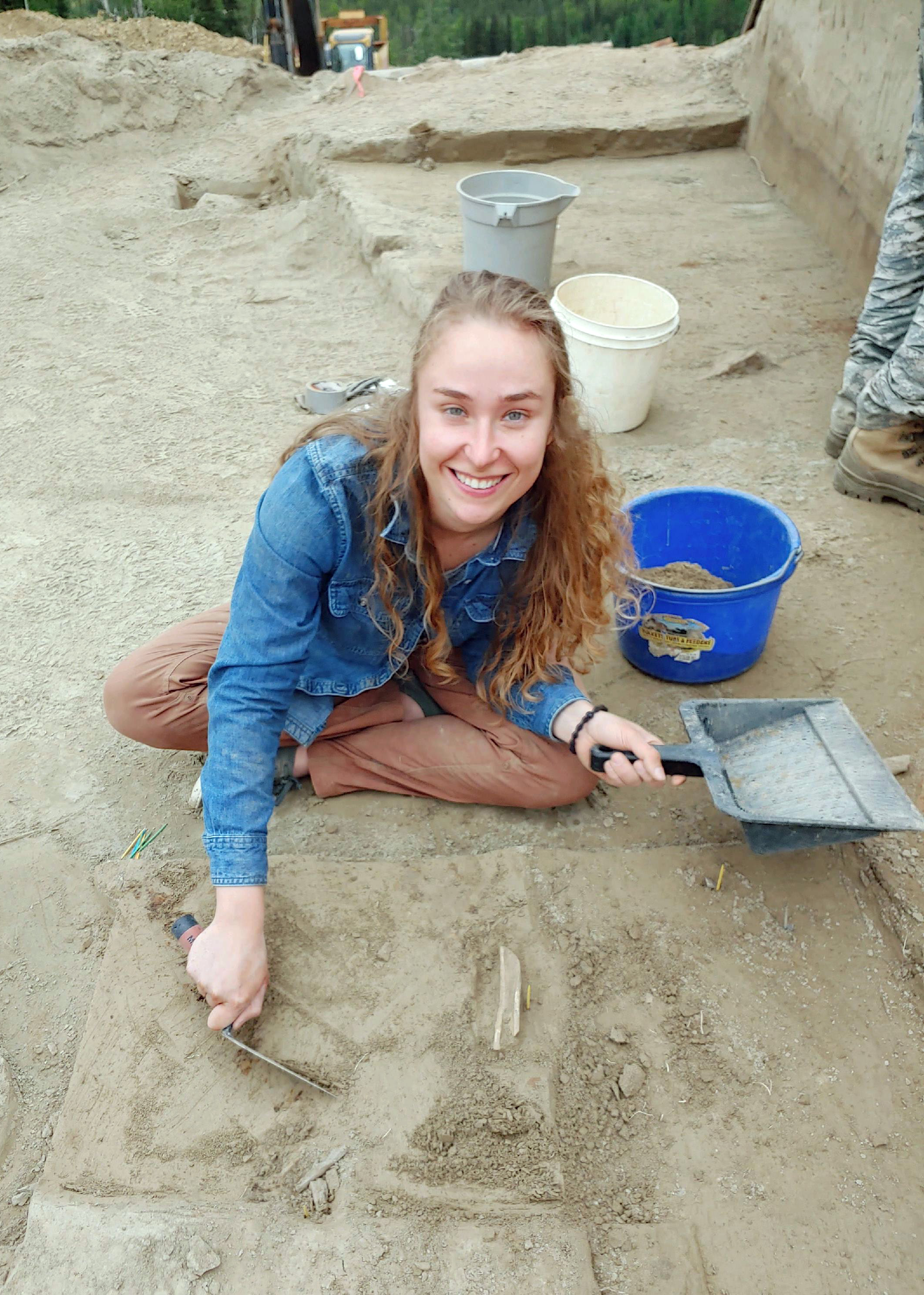 A woman holds a trowel and a dustpan while sitting on a flat dirt surface.   