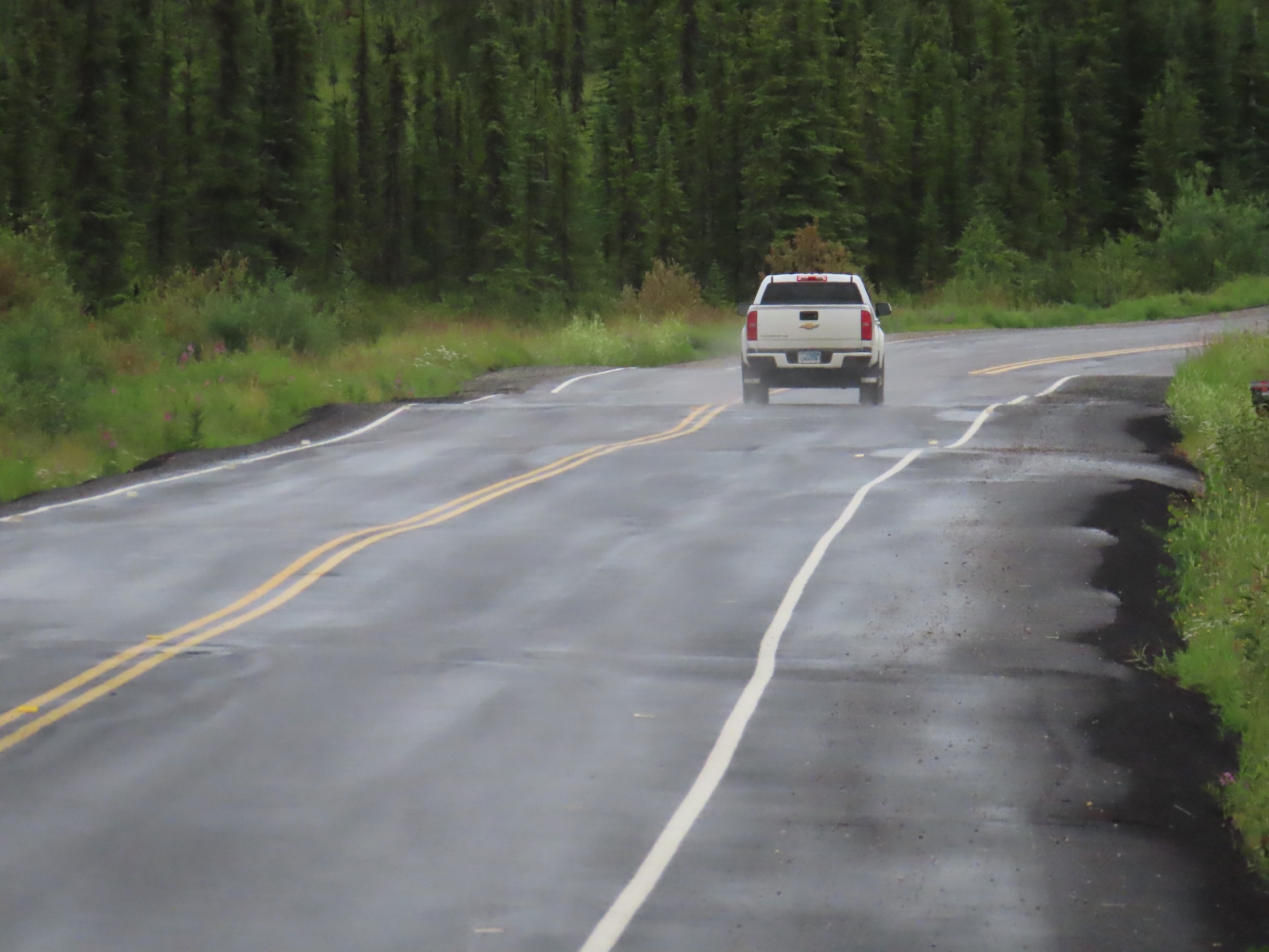 A white pickup drives down a wet, bumpy, paved and striped road with green vegetation to each side and a green tree-covered hillside beyond the approaching corner.